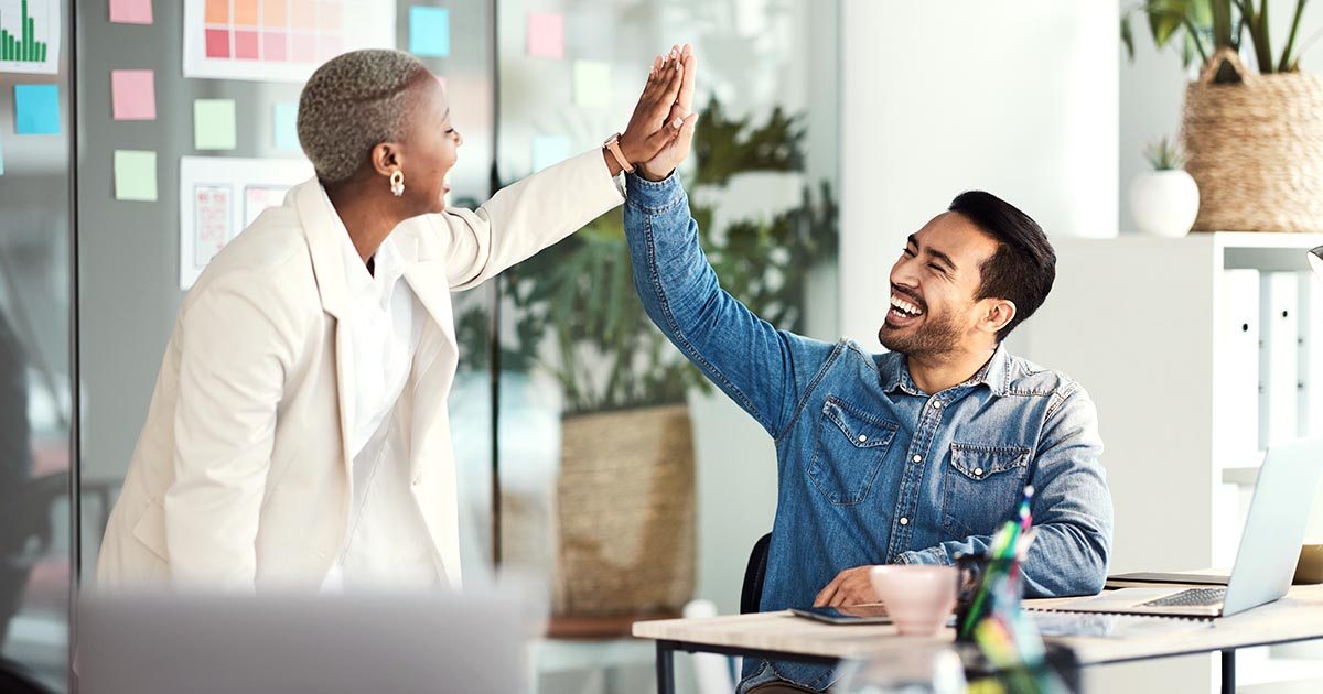 Decorative. In a modern office with glass walls, a standing woman on the left high-fives a smiling man seated at a desk on the right.