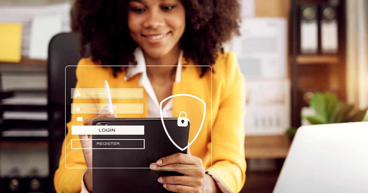Illustrative. A woman in an office smiles at her computer screen while seated at a desk facing the viewer. Superimposed over her is a graphic of a padlock and rectangles for login fields.