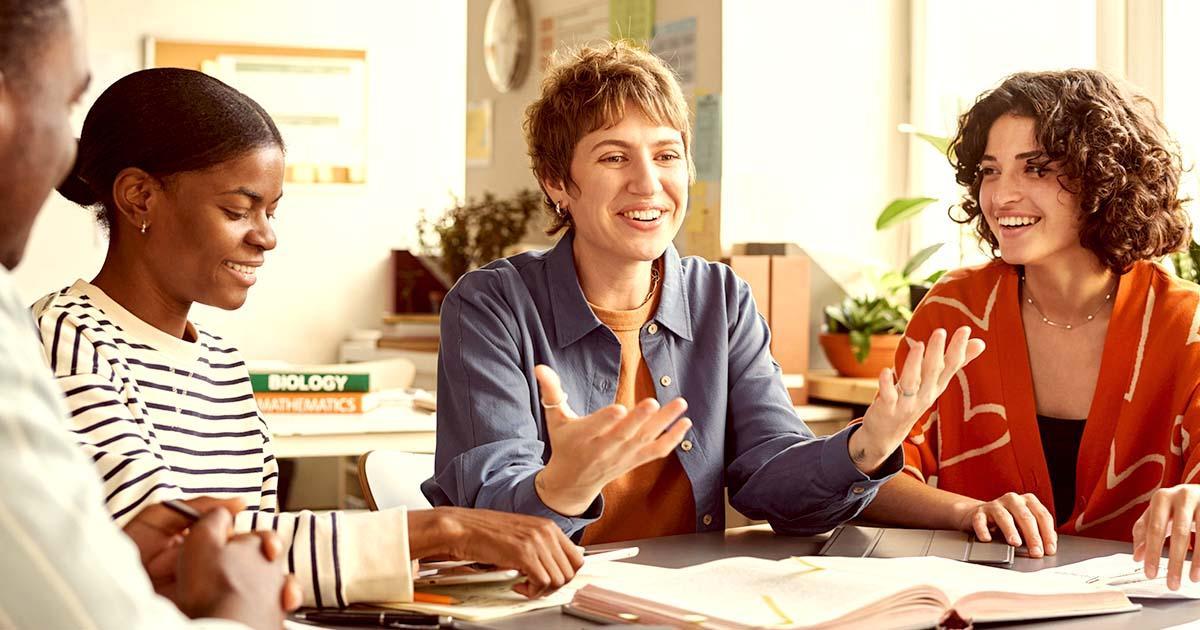 Illustrative. A group of teachers conversing in a classroom while late afternoon sunlight stream in through large windows.