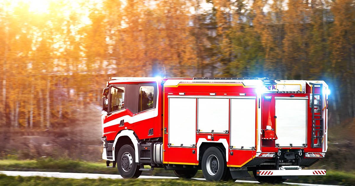 Illustrative. A firetruck speeds along a road toward the right side of the image. Sunlight streams through trees with autumn leaves in the background.