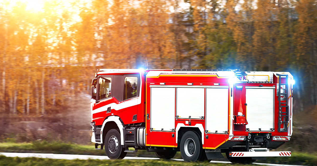 Illustrative. A firetruck speeds along a road toward the right side of the image. Sunlight streams through trees with autumn leaves in the background.