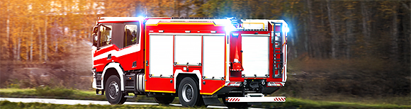 Illustrative. A firetruck speeds along a road toward the right side of the image. Sunlight streams through trees with autumn leaves in the background.