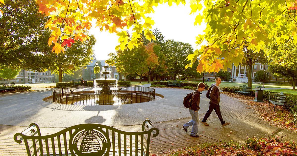 Illustrative. A photo of the iconic fountain at Radford University in Radford, Virginia. Late afternoon sunlight illuminates autumn leaves on the trees as two students walk toward the right side of the image.