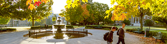 Illustrative. A photo of the iconic fountain at Radford University in Radford, Virginia. Late afternoon sunlight illuminates autumn leaves on the trees as two students walk toward the right side of the image.
