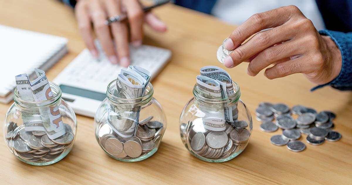 A pair of hands enters the frame from above, a coin held in the left hand. A table surface with three spherical, clear glass jars fills the rest of the frame. The jars are filled with varying amounts of cash. On the left of the frame is a partial view of a notepad of paper under the right hand.