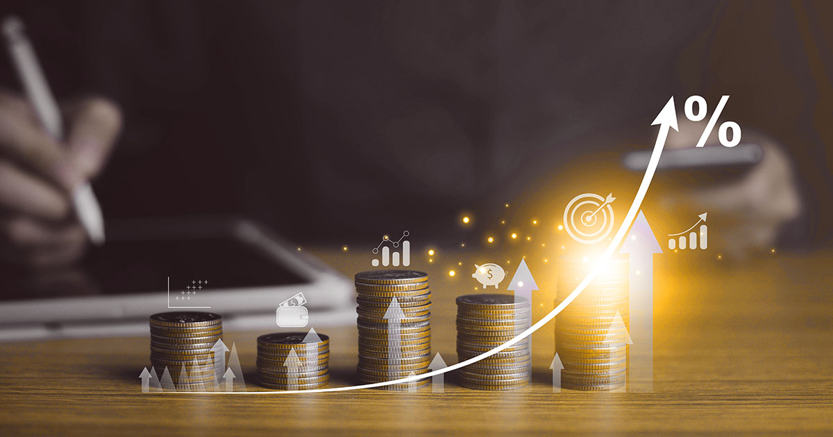 Decorative. Five stacks of coins on a tabletop with a curved graphic over them indication increase. In the background on the left is a hand using a stylus on a computer tablet. To the right of the coins is a vase of sunflowers.