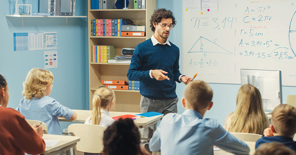 Decorative. A young teacher lecturing students in his classroom. A large whiteboard and a bookcase are on the wall behind him.