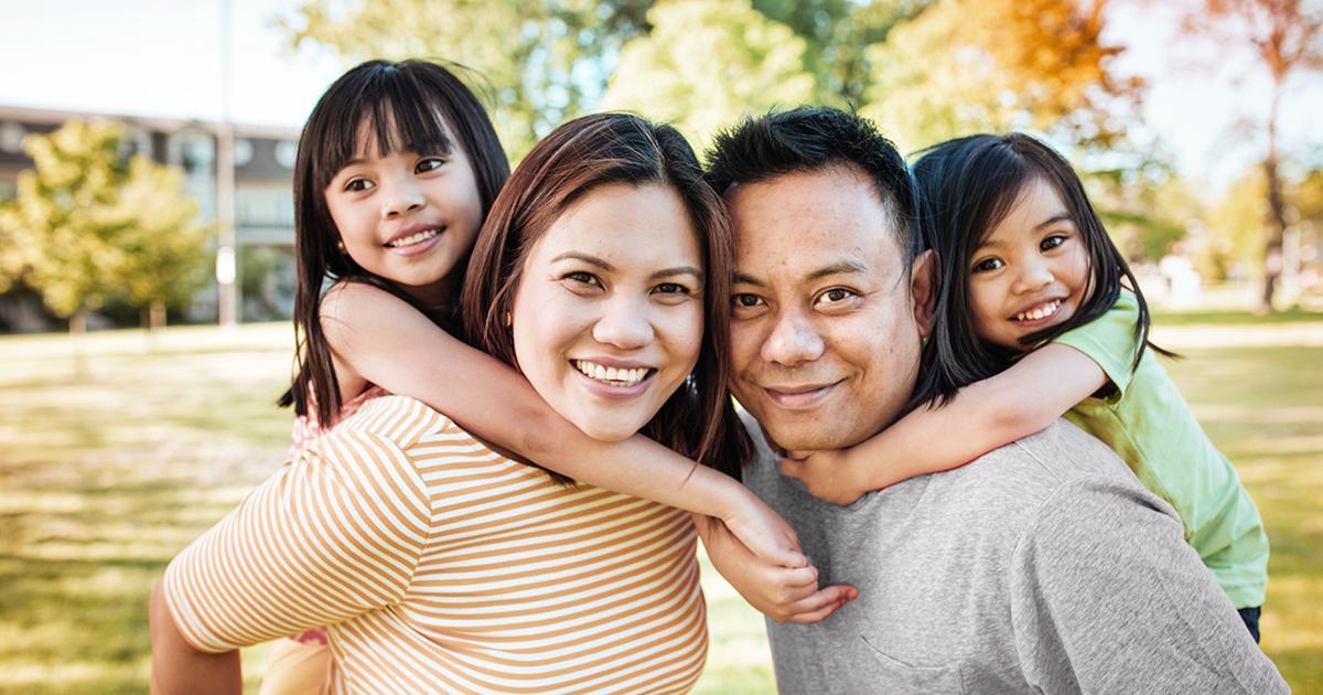 Decorative. A family of four embraces and smiles for the camera, posing in a park like setting.