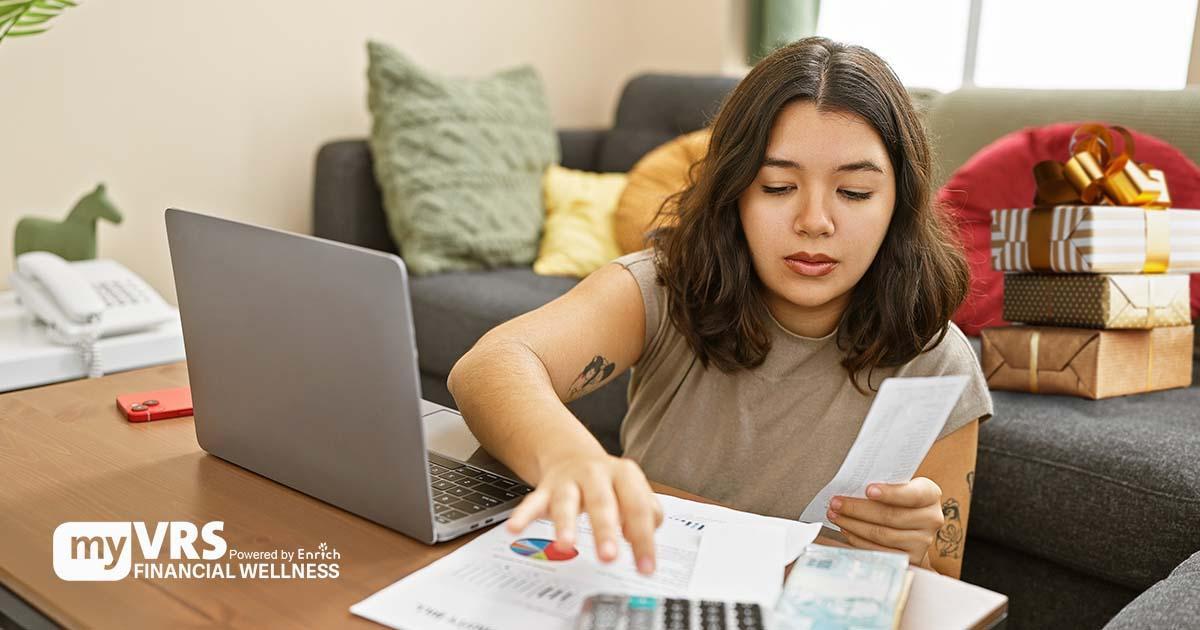 Illustrative. A young woman seated on the floor of her living room works on her budget with papers and a laptop computer in front of her on a low table. Behind her are wrapped gifts on an upholstered couch.