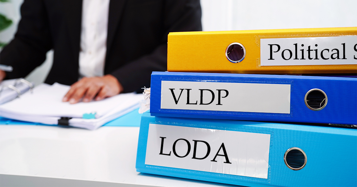 Decorative. On the right, three loose leaf binders are stacked on a table. Labels on their spines read political subdivisions, VLDP and LODA. in the bckground, on the right, a person in professional dress sits on the opposite side of the table, working on some papers.