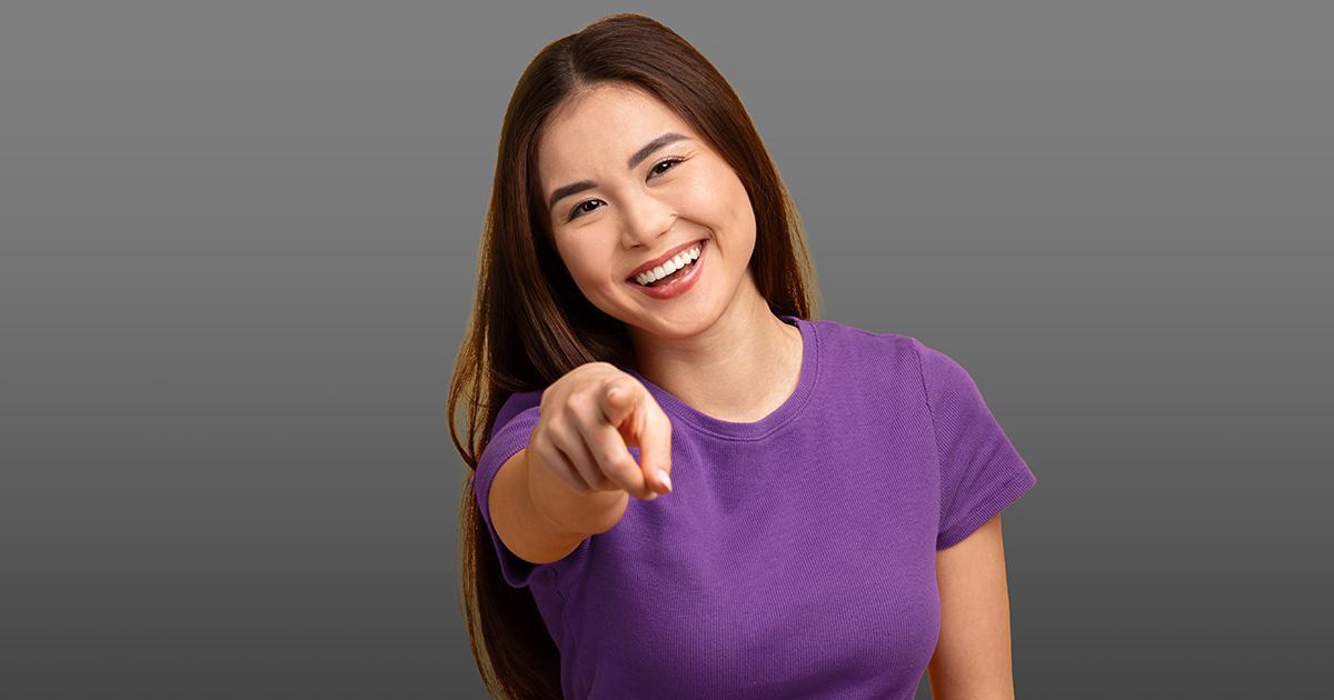 A smiling woman with long hair, wearing a short sleeved shirt, points directly at the viewer.