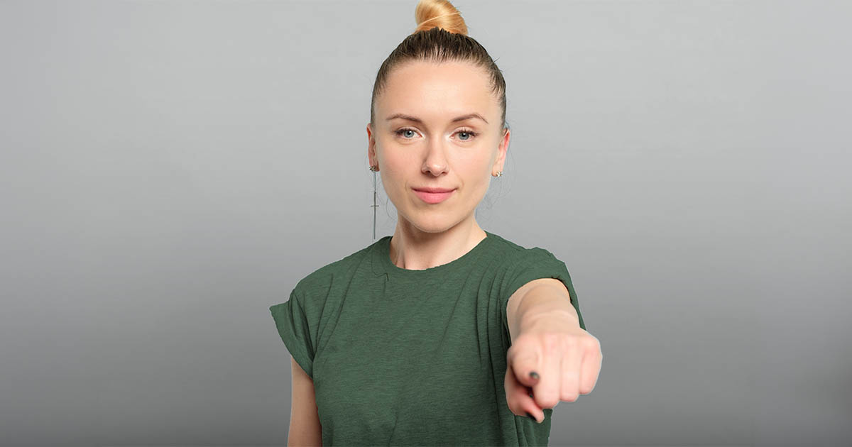 Decorative. A young woman with a closed-lip smile points at the viewer.