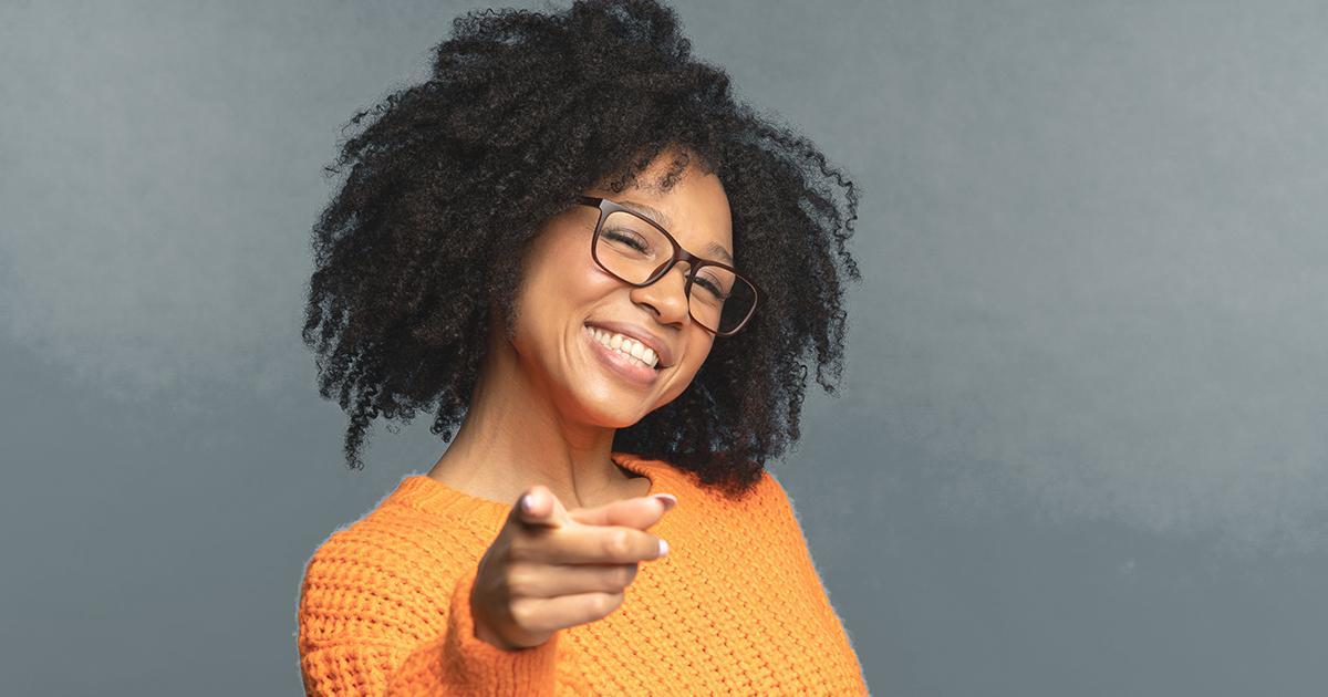 Decorative. A woman tilts her head and smiles broadly at the viewer while pointing at the viewer with her right index finger.