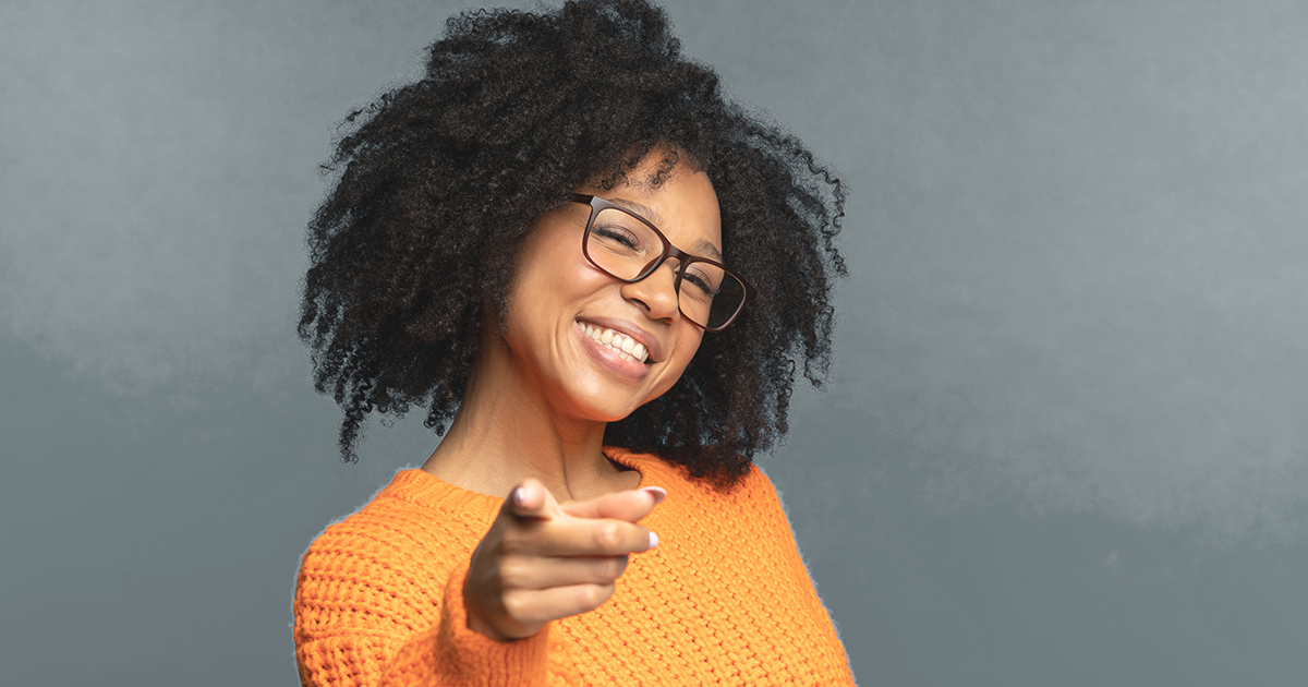 Decorative. A woman tilts her head and smiles broadly while pointing at the viewer with her right index finger.