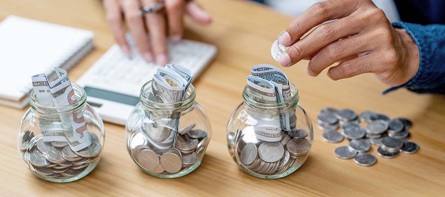 Coins sorted into glass jars on a table while hands organize additional coins nearby, illustrating savings or financial planning.