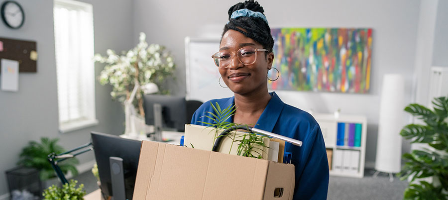 Photograph of a person holding a cardboard box filled with office supplies, standing in a modern office setting with desks, computers, and colorful artwork on the wall. The scene suggests a workplace transition or moving out, highlighted by the packed box containing a plant, files, and personal items.