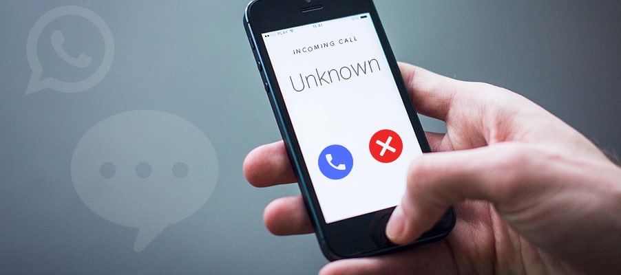 Photograph of a hand holding a smartphone displaying an incoming call from an unknown caller. Screen shows two buttons: a blue phone icon to answer and a red X icon to decline the call, with a blurred background featuring WhatsApp chat icons.