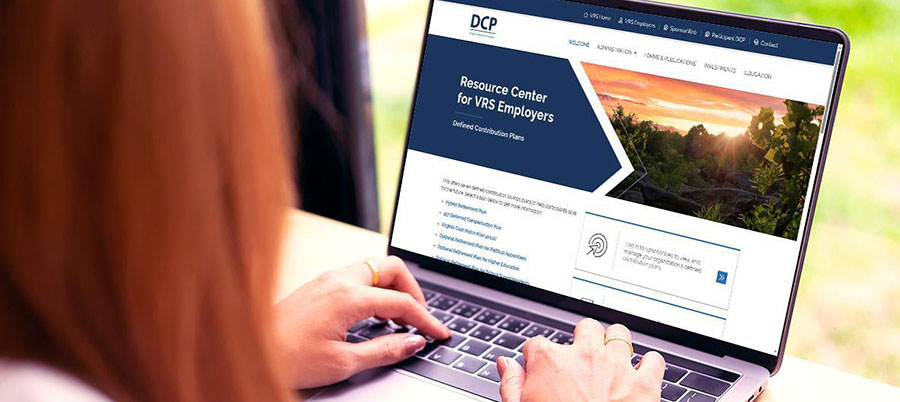 Decorative. An over-the-shoulder view of a young woman typing on a laprop computer. On the screen is the Voya Sponsorweb website. In front of the woman's laptop is a large window with a view of short vegetation.